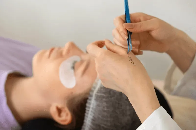 The image depicts a close-up view of a professional carefully applying eyelash extensions.