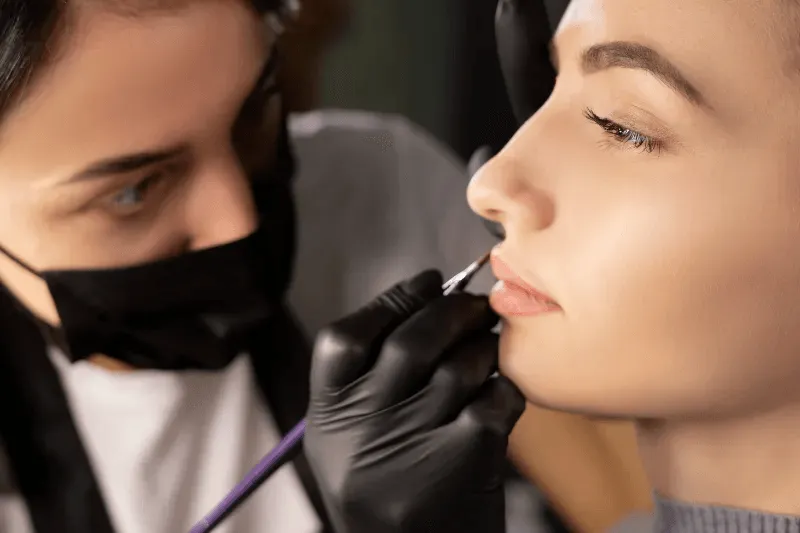 The image shows a close-up of a professional applying a lip blush tattoo, highlighting the careful and precise nature of the procedure.