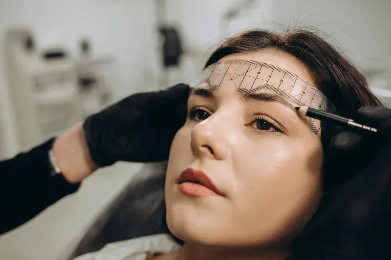 The image shows a woman having her eyebrows measured and outlined with a ruler tool in preparation for a microblading procedure