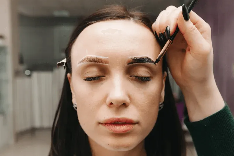 This image shows a close-up of a woman receiving eyebrow shaping, where a professional is applying a dark pigment to her brows.