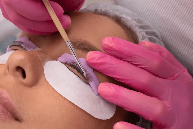 The image shows a technician carefully applying a lash lift solution to a client's eyelashes using a precision tool,