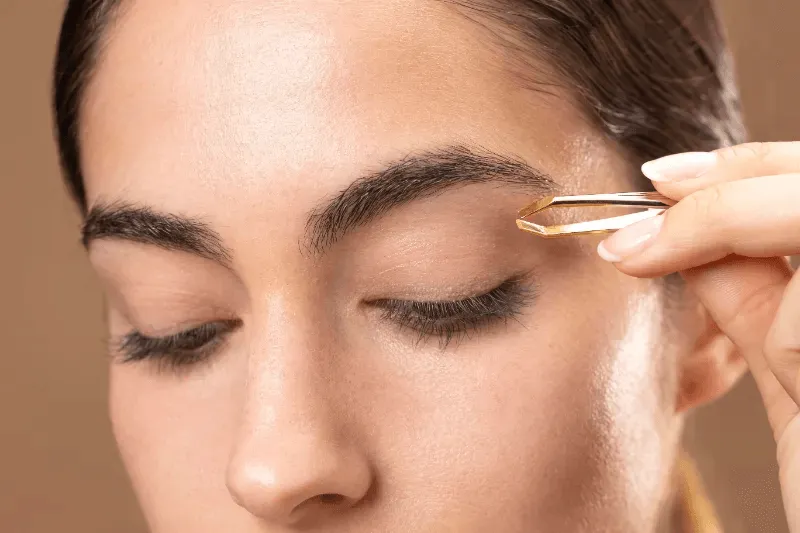 A close-up of a person shaping their brows with tweezers, highlighting post-procedure maintenance.