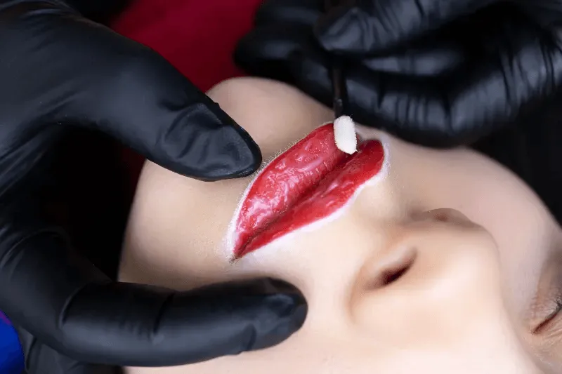 The image depicts a lipstick tattoo procedure, highlighting the precision and care involved in applying permanent lip color.